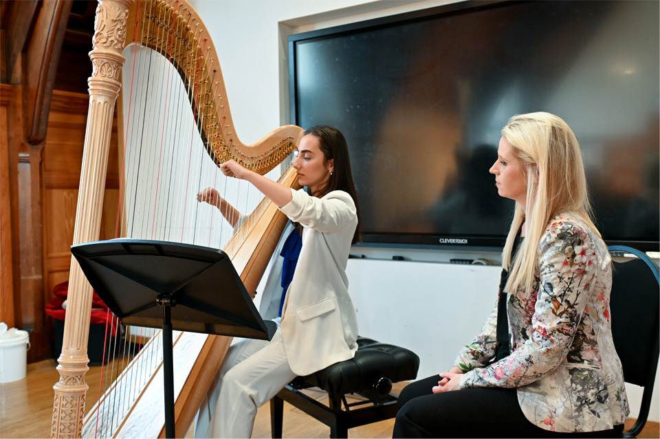 A female student, wearing a white suit, playing the harp, with a woman with blonde hair, sitting next to her observing the student.
